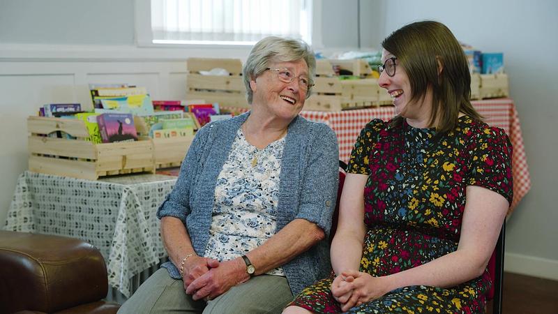Two white women sit on chairs smiling at each other, sharing a joke
